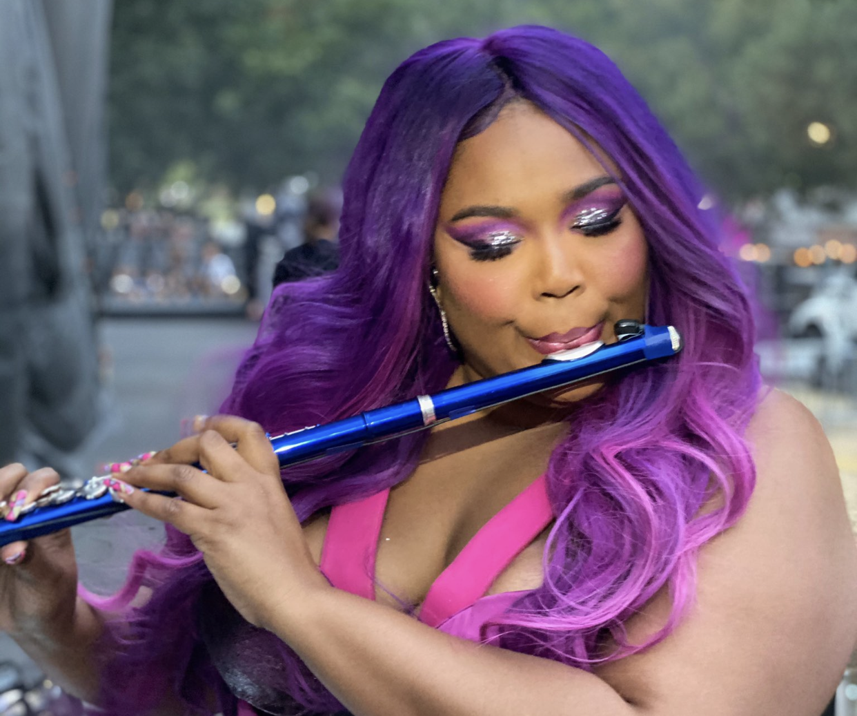 Lizzo playing a blue wind instrument with her purple hair and light purple highlights.