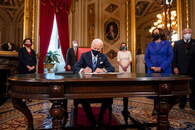 Joe Biden signing documents with Kamala Harris in the background.
