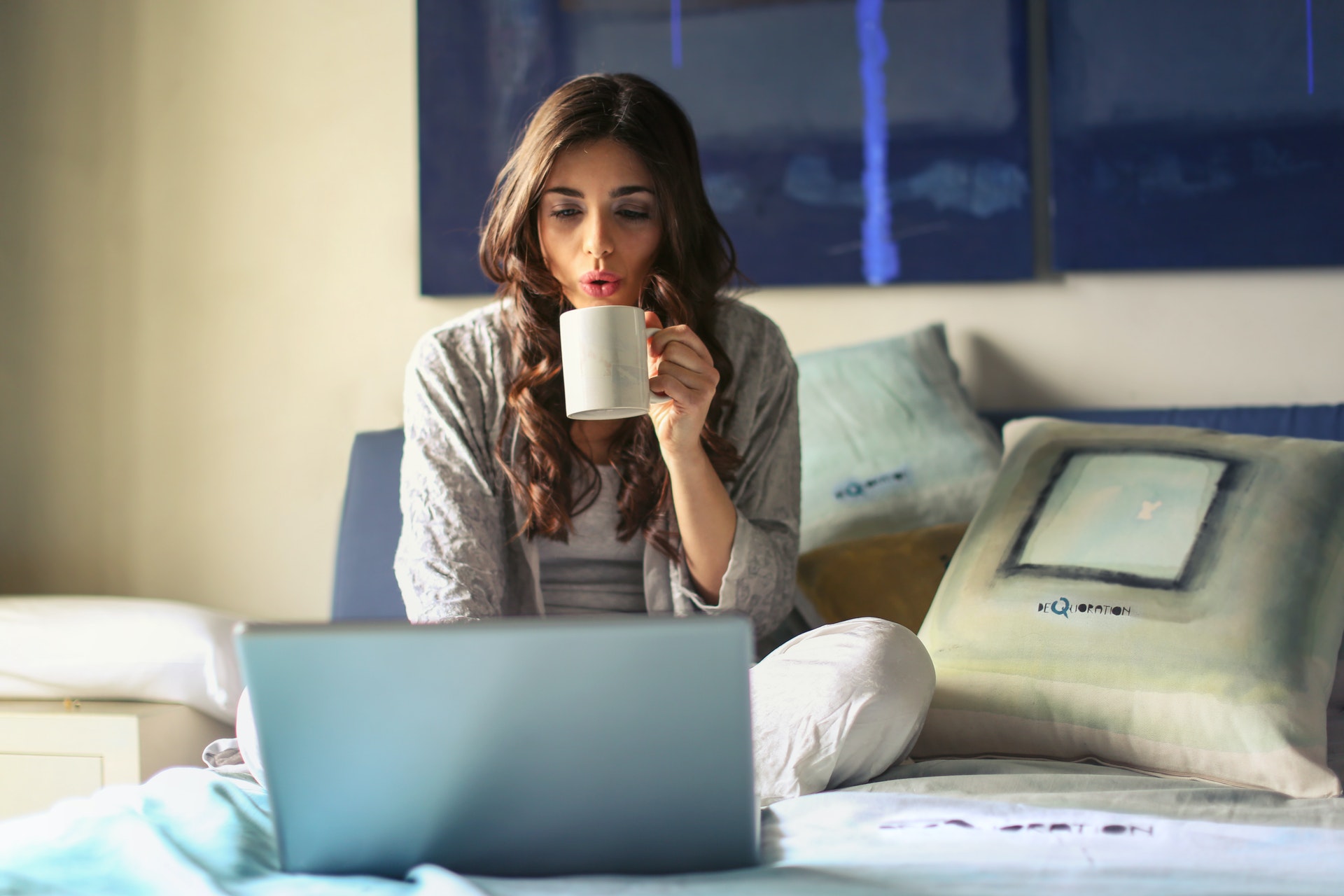 A woman working from their bed drinking coffee looking at a laptop